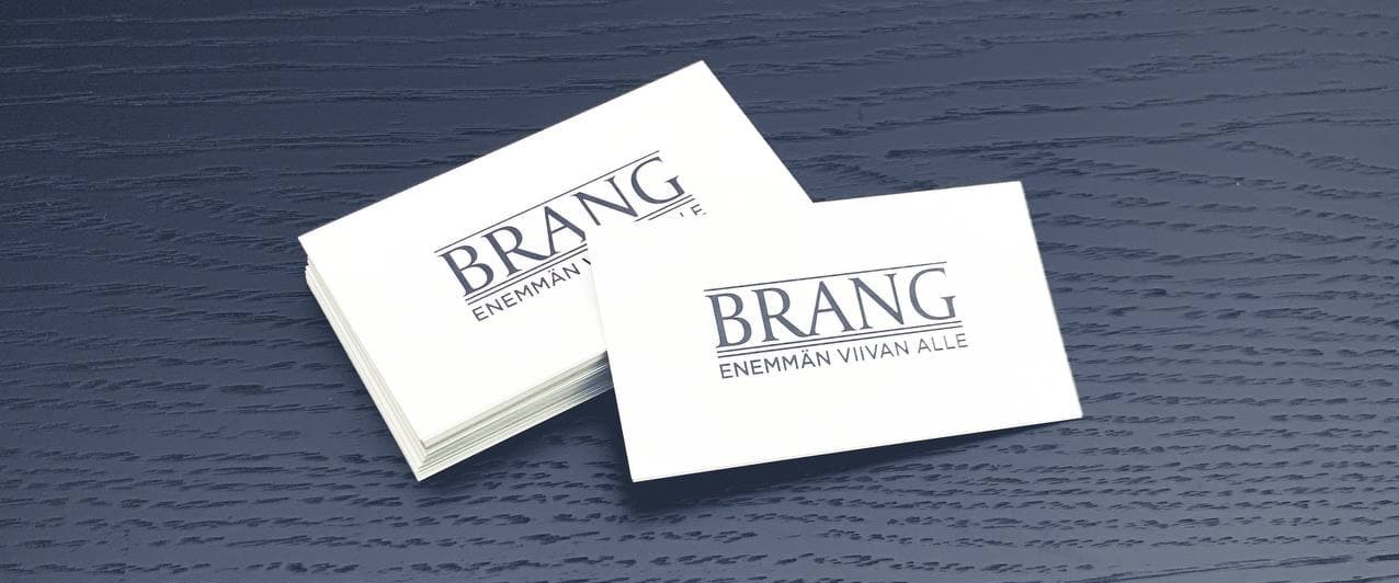 Kayntikortit Kaksi pinoa valkoisia käyntikortteja tummansinisellä puupinnalla. Käyntikorteissa on Brang-logo ja teksti "Enemmän viivan alle".